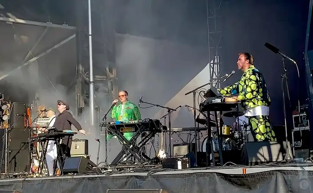 Musicians performing on stage, one in a green outfit with circles, another in a black outfit and hat.