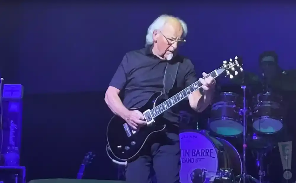 Musician with white hair playing a black electric guitar on stage with a drum set in the background.