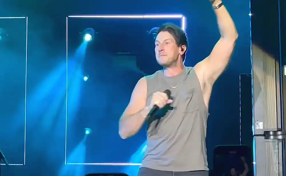 A male singer with arms raised, holding a microphone and wearing a grey tank top on a blue-lit stage.