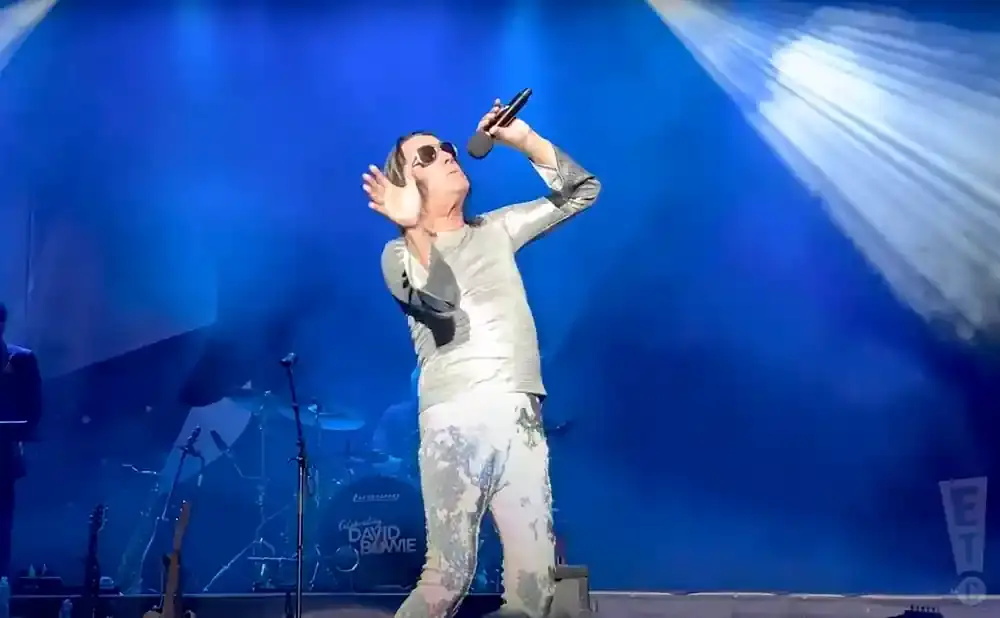 A singer in a silver outfit and sunglasses performs on a blue-lit stage, holding a microphone.
