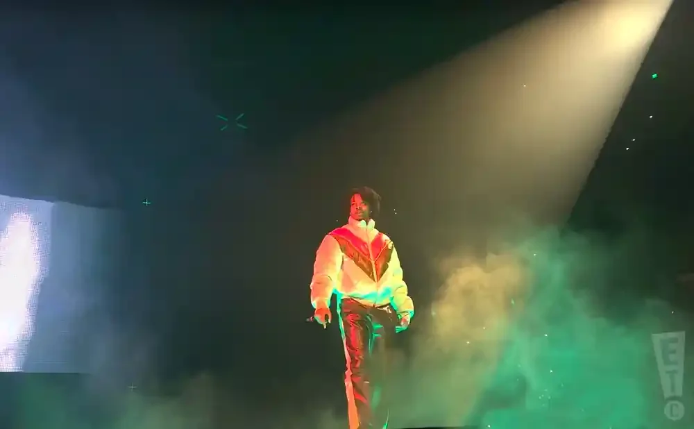 A person stands on a dimly lit stage, illuminated by a spotlight, wearing a white jacket with red and orange chevron stripes and shiny black pants.