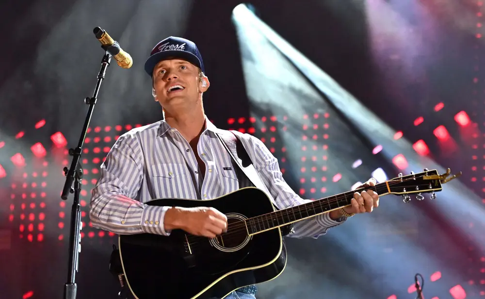 A man in a striped shirt and baseball cap plays an acoustic guitar on stage with red lights in the background.