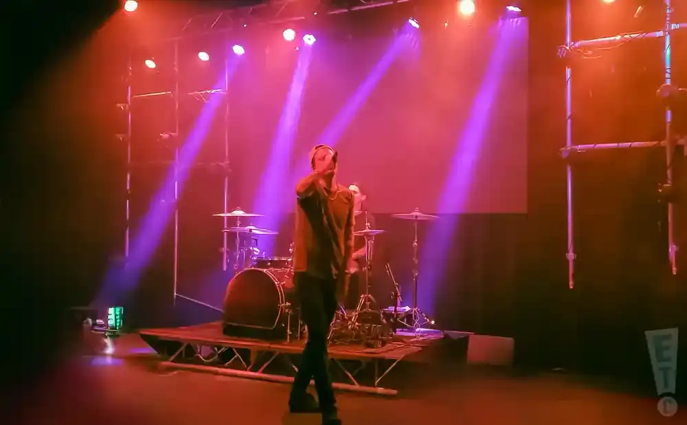 A male singer performs on stage with a drum set in the background, illuminated by vibrant pink and red stage lights.