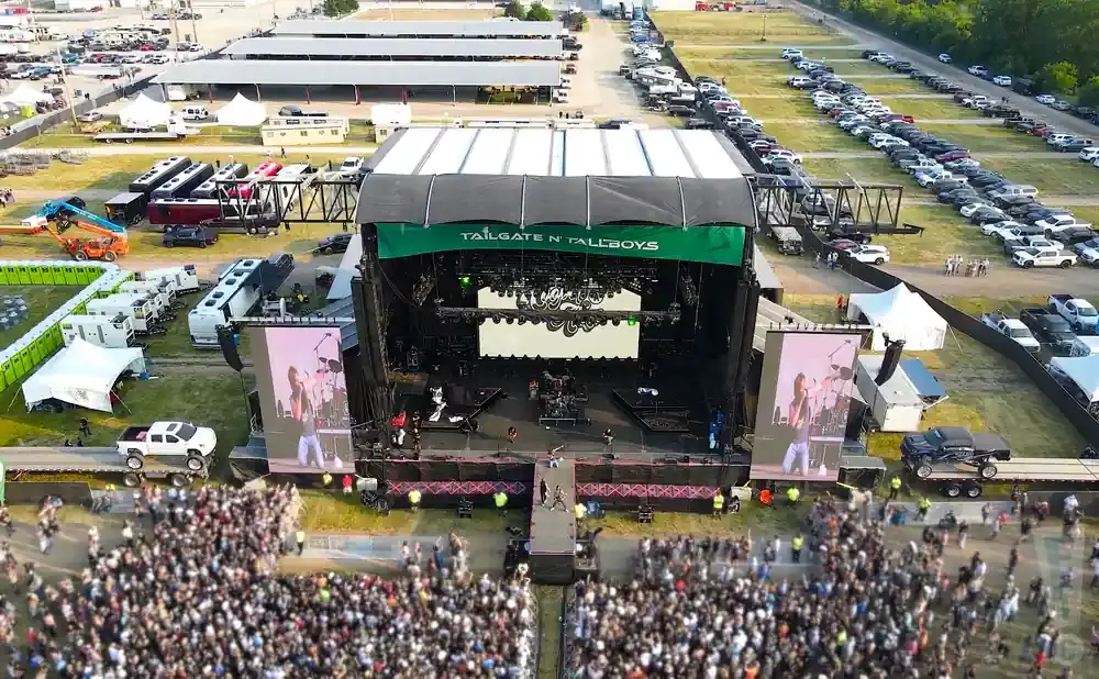 Aerial view of an outdoor concert stage with a large crowd, trucks, and RVs.