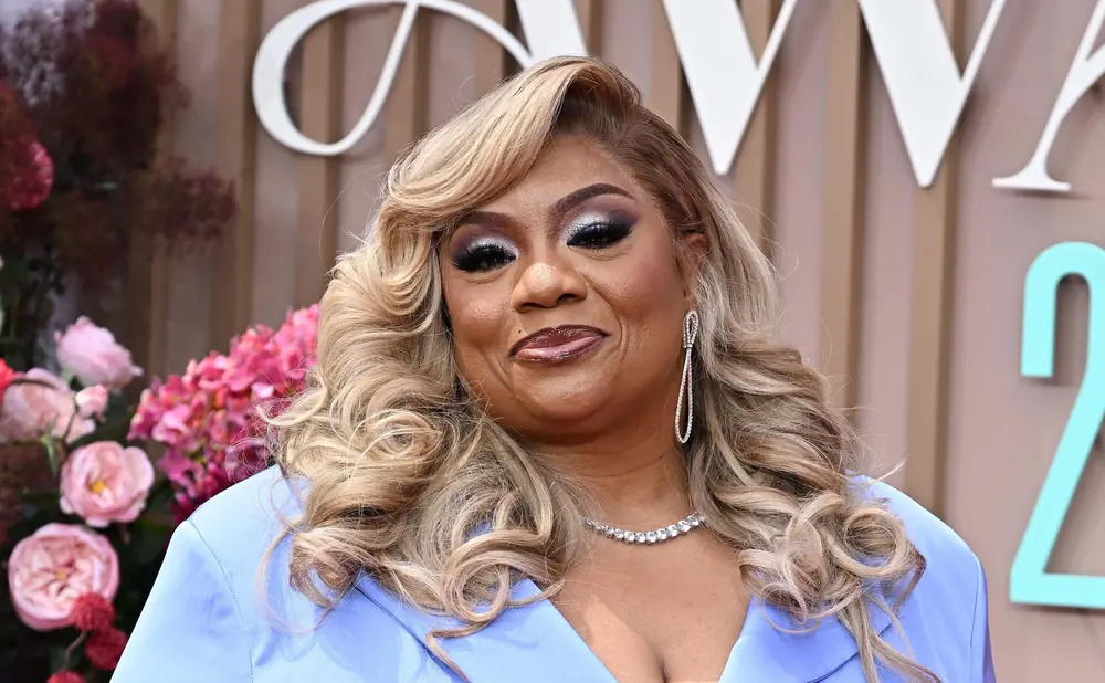 A woman with blonde curly hair, heavy makeup, and a blue dress smiles.