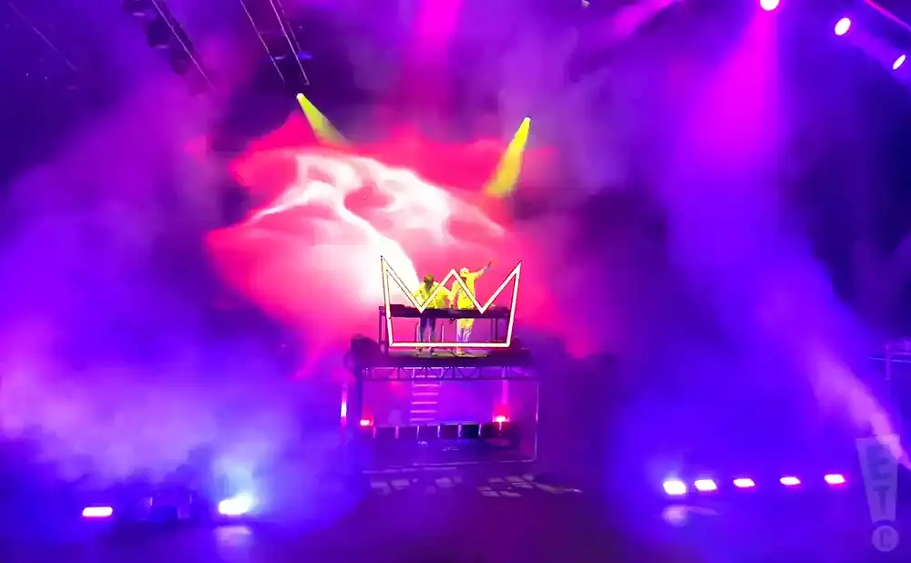 Two DJs in neon yellow outfits perform on a stage with a giant neon crown and colorful smoke.