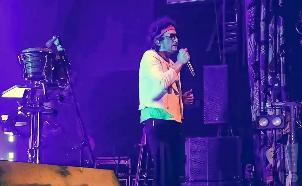 A man in a yellow jacket and red headband sings into a microphone on a purple-lit stage.