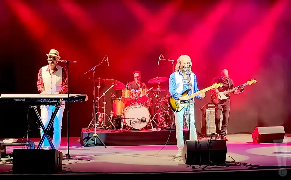 A band performs on a stage with red lighting: a keyboardist, drummer, guitarist, and bassist.