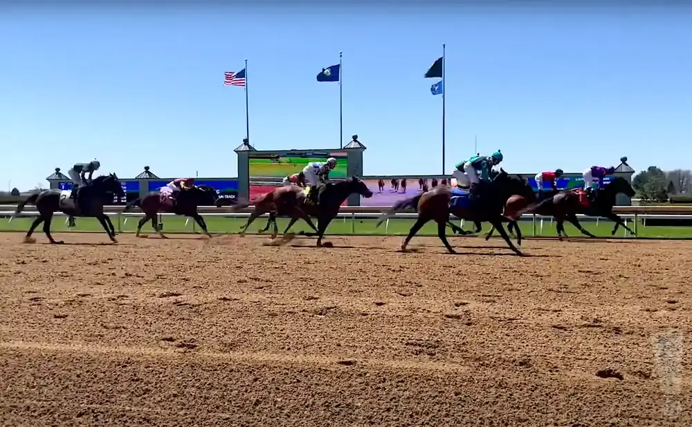 Horses and jockeys race on a dirt track under a clear blue sky, with flags and a large screen in the background.