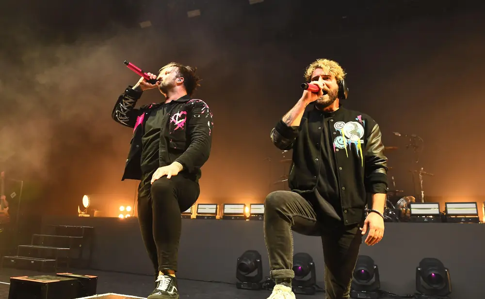 Two male singers perform on stage in black jackets, with spotlights and smoke in the background.