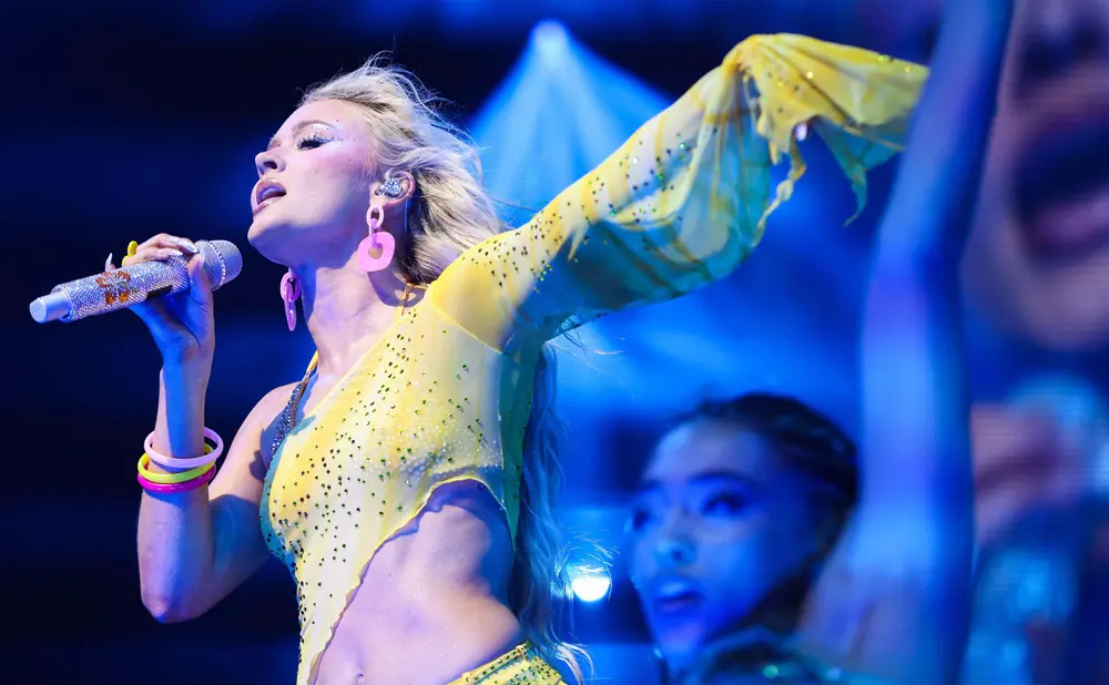 A singer in a sparkly yellow outfit sings into a bejeweled microphone, with a dancer blurred in the background.