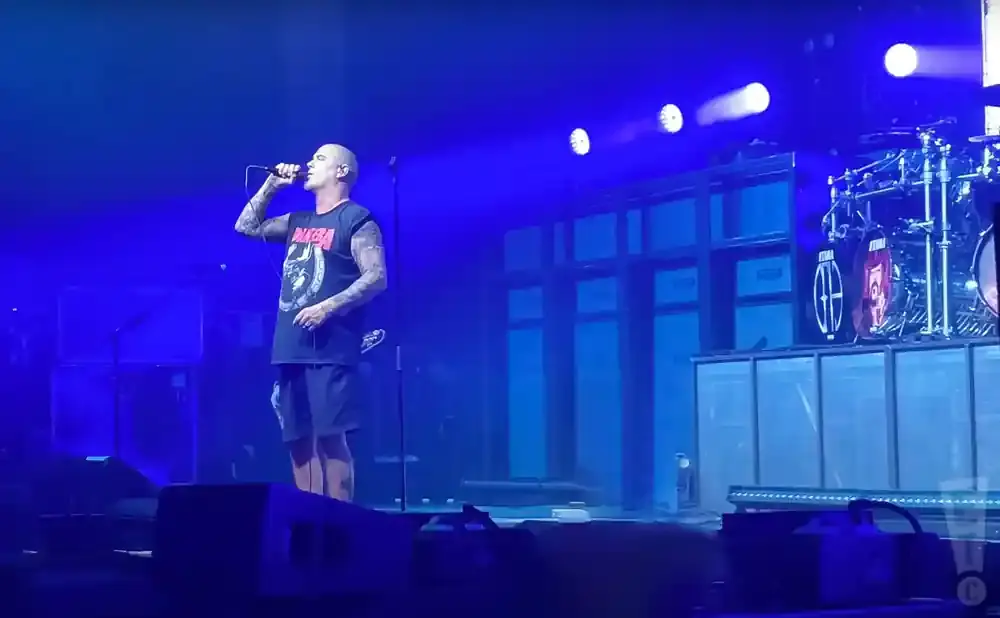 A bald man with tattoos on his arms sings into a microphone on a stage with blue lighting and a drum set.
