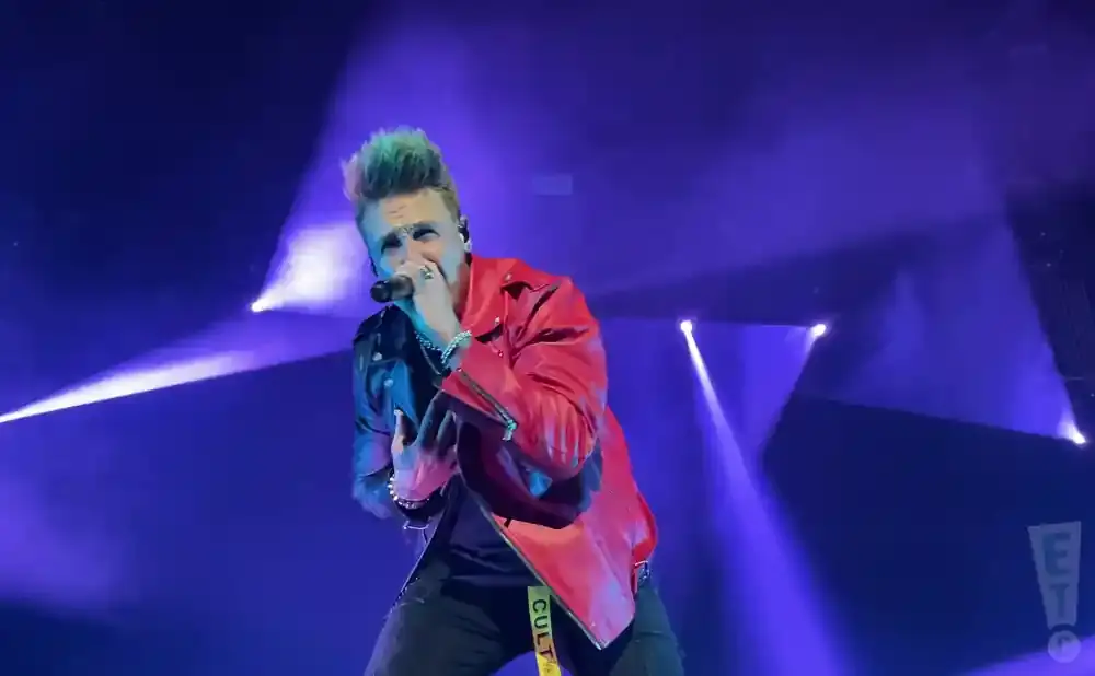 A man with spiky green hair and a red jacket sings into a microphone on a purple-lit stage.