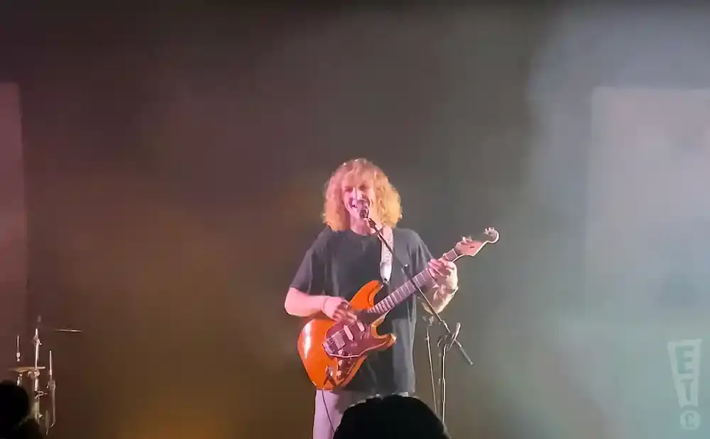 A blonde guitarist with a bright orange electric guitar smiles as they play on stage with a hazy background.