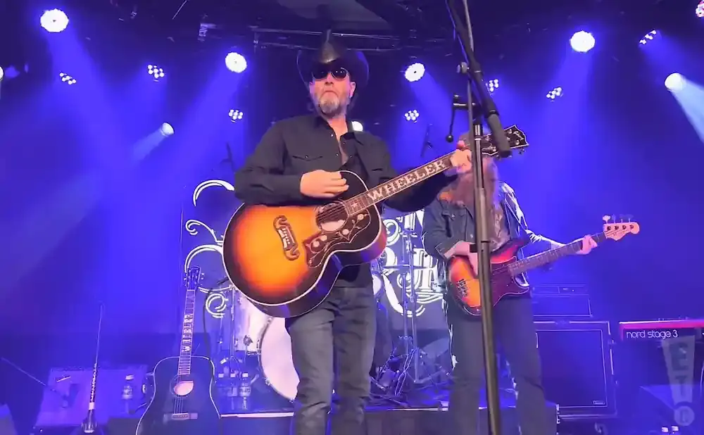 Man in cowboy hat plays acoustic guitar on stage with blue lights.