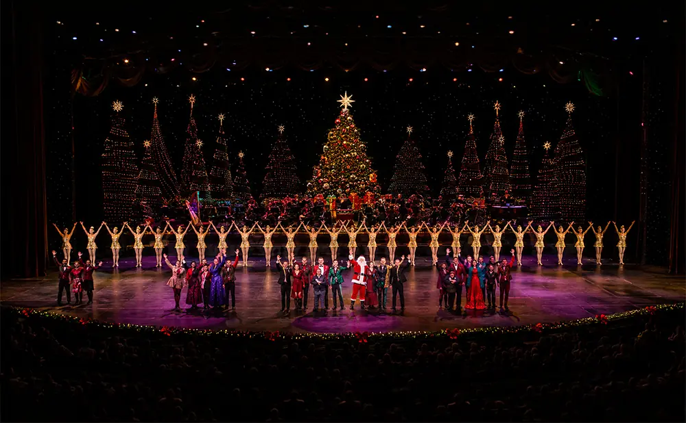A stage show with dancers, singers, and Santa Claus under a lit Christmas tree and backdrop of illuminated trees.
