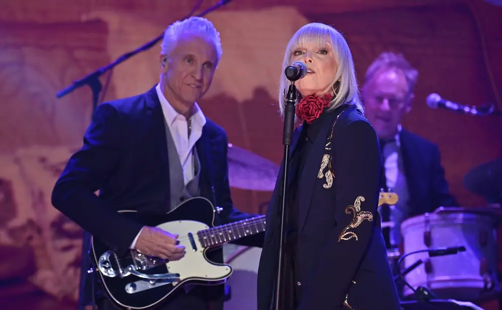 Singer with silver hair performs on stage, wearing a dark jacket with gold embroidery, while a guitarist plays beside her.