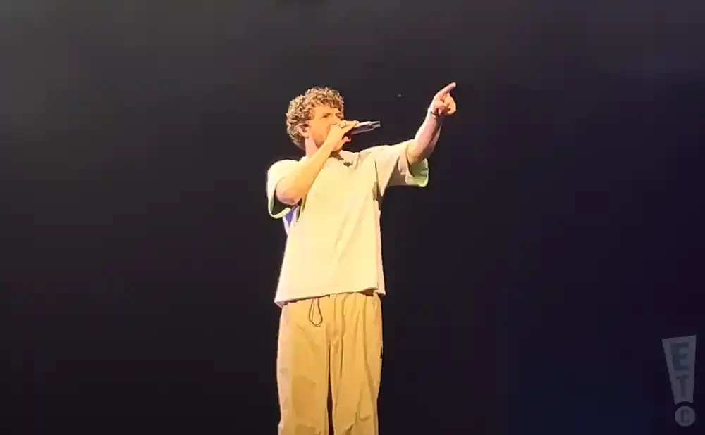 A man with curly hair, wearing a light-colored t-shirt and tan pants, sings into a microphone on a dark stage.