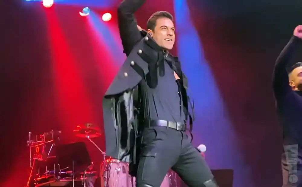A male performer in a black military-style jacket and pants on stage with red and blue spotlights.