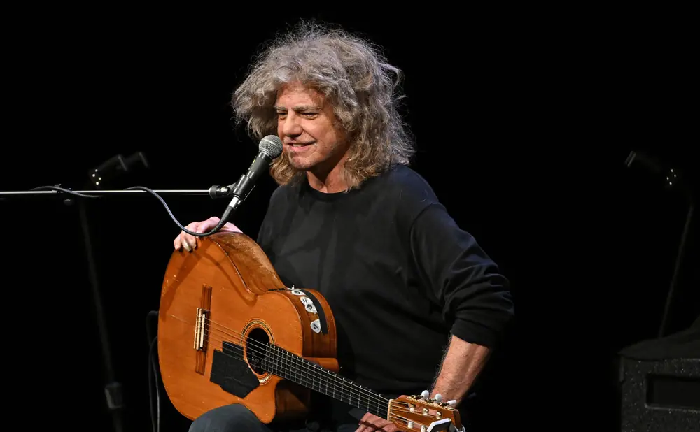 A man with wild, curly grey hair plays an acoustic guitar on stage.