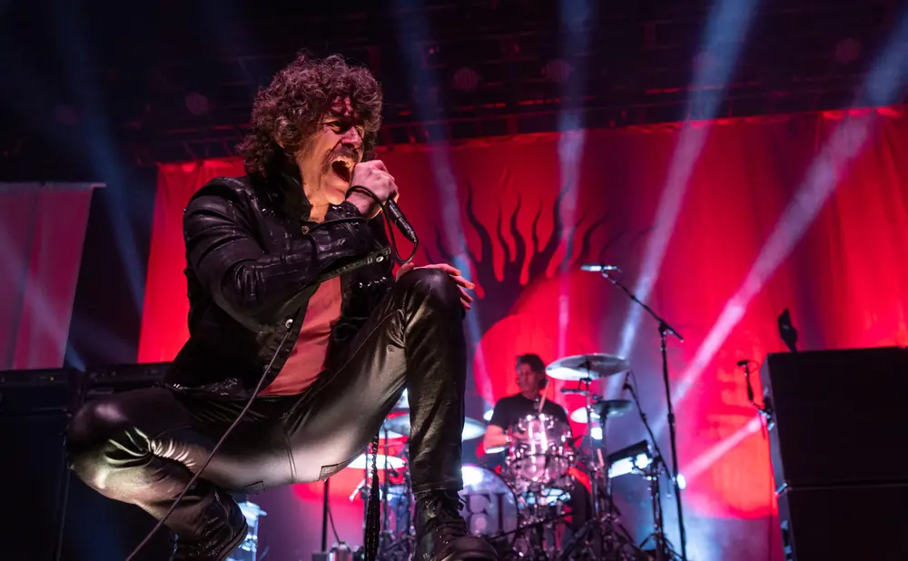 A male rock singer with curly hair and a mustache crouches and sings into a microphone on a stage with red lighting.