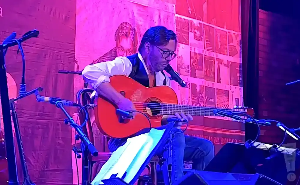 A guitarist plays an orange acoustic guitar on stage under pink and blue lighting.