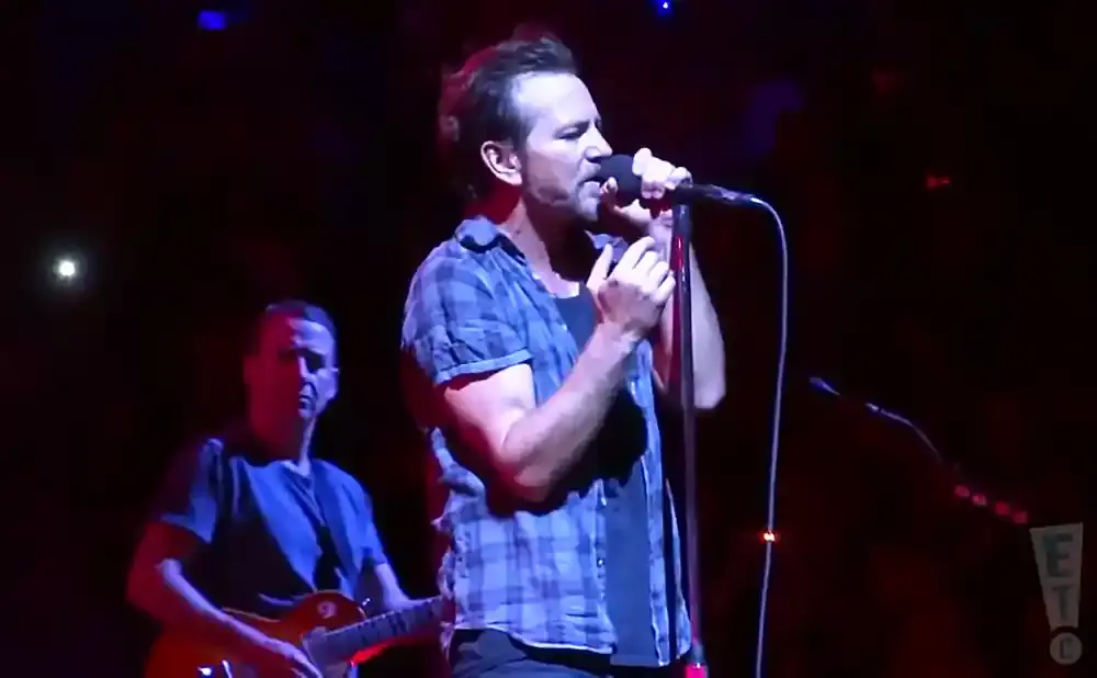 A man in a blue plaid shirt sings into a microphone on stage, with another man playing guitar behind him.