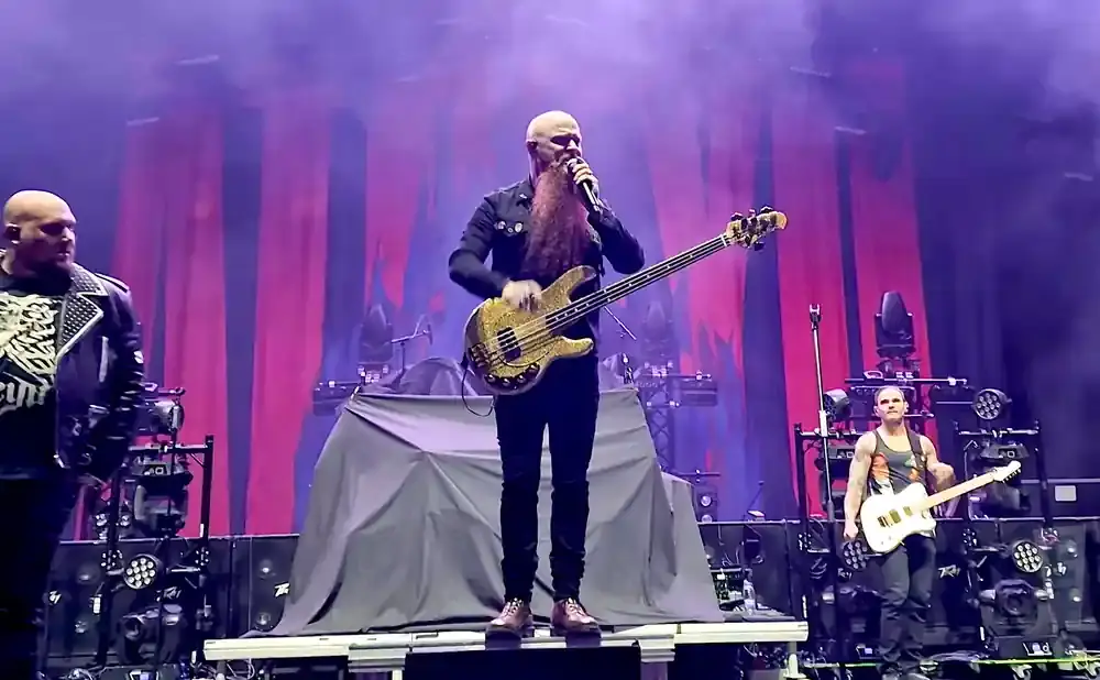 A bald bassist with a long beard plays a gold bass guitar on stage, with two other men standing beside him.