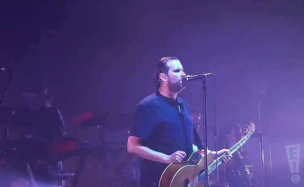 A man sings into a microphone and plays an acoustic guitar on a stage with purple lighting.