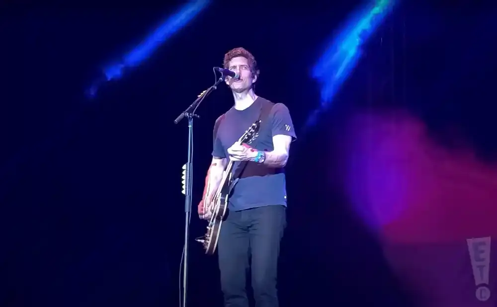A male guitarist plays his electric guitar on stage under blue and pink spotlights.