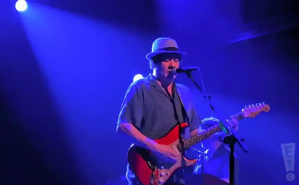 A man in a hat plays a red electric guitar on a blue-lit stage.