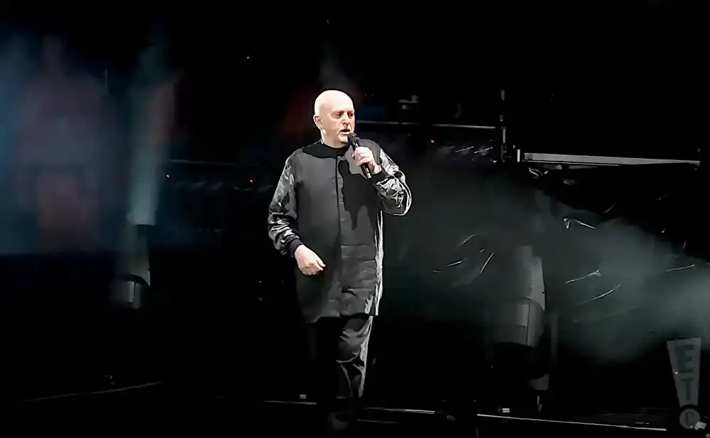 A bald man in a dark, patterned jumpsuit holds a microphone on a dimly lit stage.