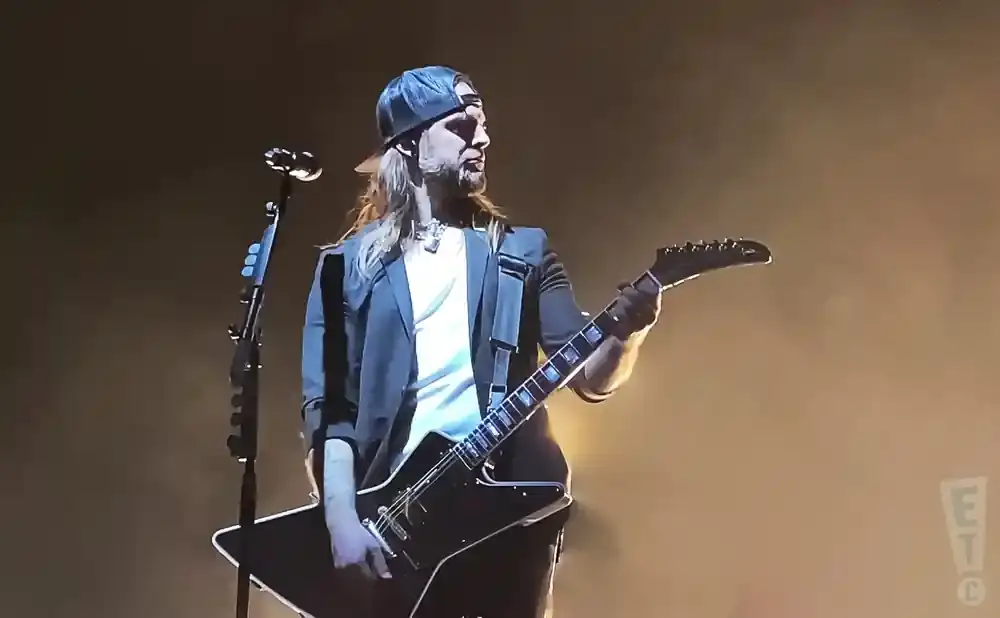 Guitarist with long blonde hair, beard, and baseball cap plays a black electric guitar on stage.