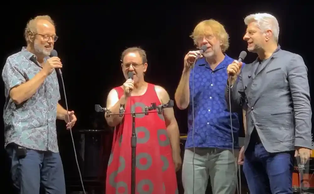 Four men on stage singing into microphones, two in casual shirts and two in more formal attire.