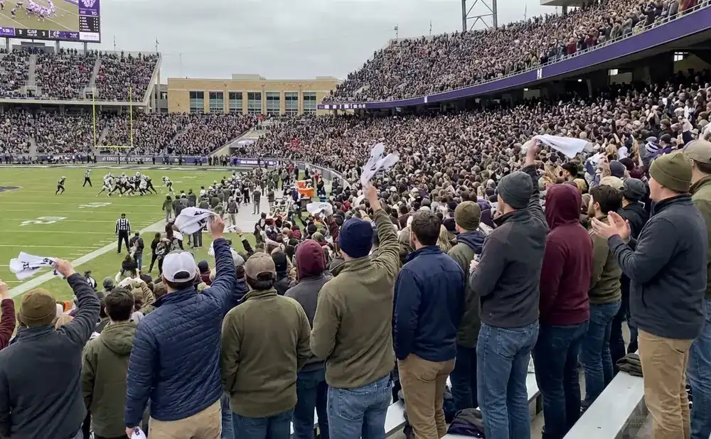 Fans in a football stadium wave white towels as players face off on the field under a cloudy sky.