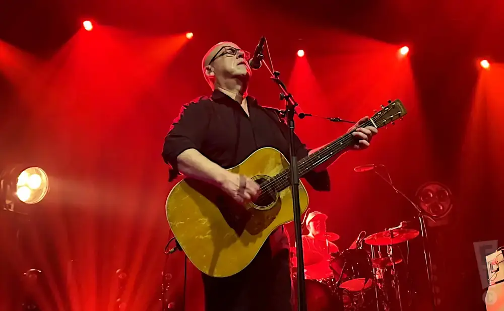 A bald man in glasses plays an acoustic guitar on a brightly lit red stage.