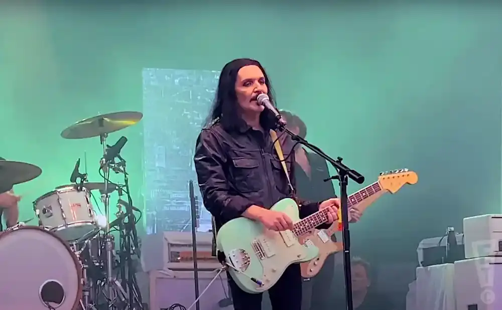 A pale-skinned musician with long black hair and heavy eyeliner plays a light blue electric guitar and sings into a microphone on stage.