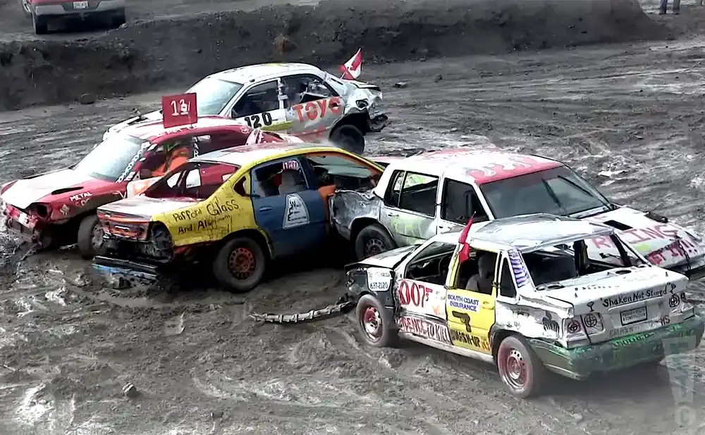 Demolition derby cars are mud-splattered and damaged. A yellow and blue car is center.