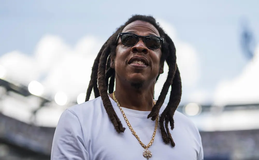 Jay-Z in sunglasses and a white t-shirt with a gold chain, outdoors.
