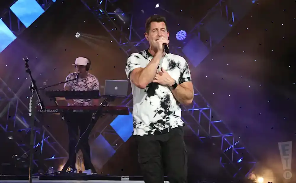 Singer on stage with a keyboard player, both wearing tie-dye shirts, with dramatic lighting.
