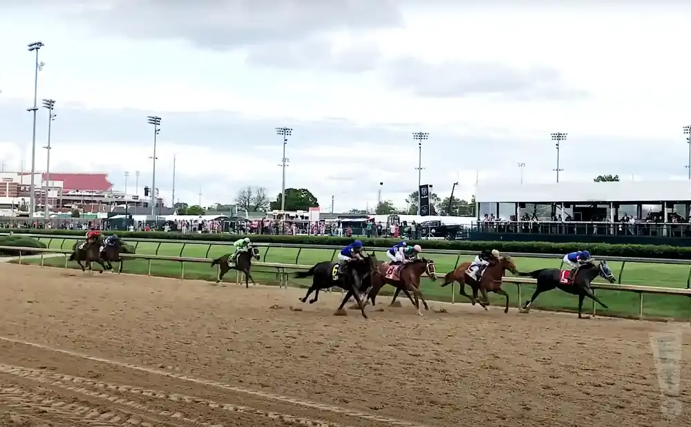 Horse race with multiple horses and jockeys on a dirt track, with grandstands and lights in the background.