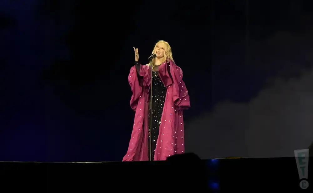 Singer in a sparkling black dress and a flowing pink robe performs on stage.