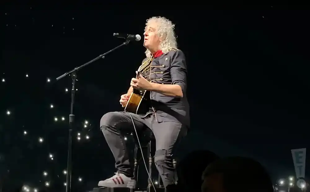 A man with curly blonde hair, playing an acoustic guitar and singing into a microphone while sitting on a stool on a dark stage with many small bright l…