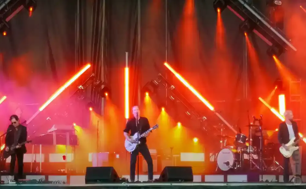 A band performs on a stage lit by bright orange lights, with guitars and drums visible.