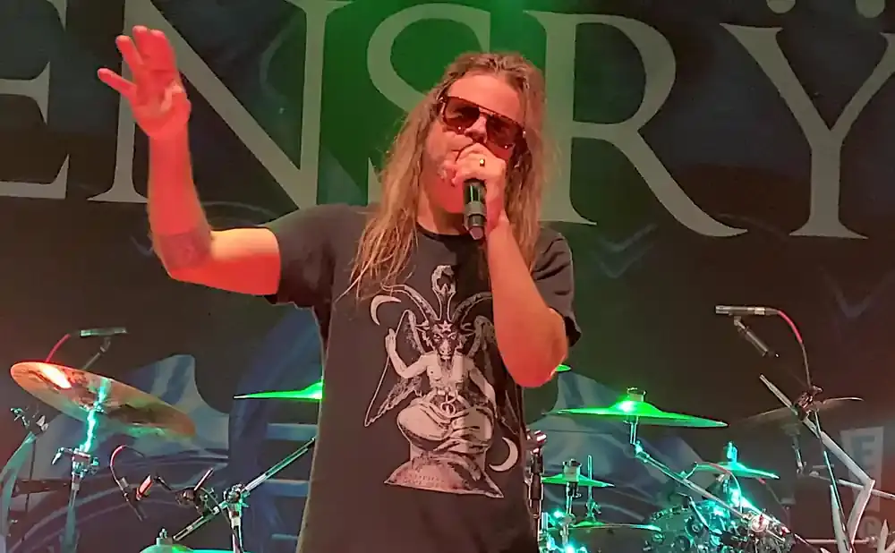A male singer with long hair and sunglasses sings into a microphone, wearing a t-shirt with a Baphomet image.