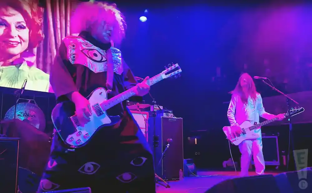 A band performs on stage under purple and blue lights. A guitarist in a colorful, eye-patterned outfit plays a white guitar.