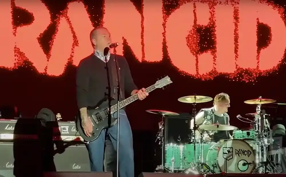 A punk band performs on stage, with the lead singer playing guitar in front of a large red 