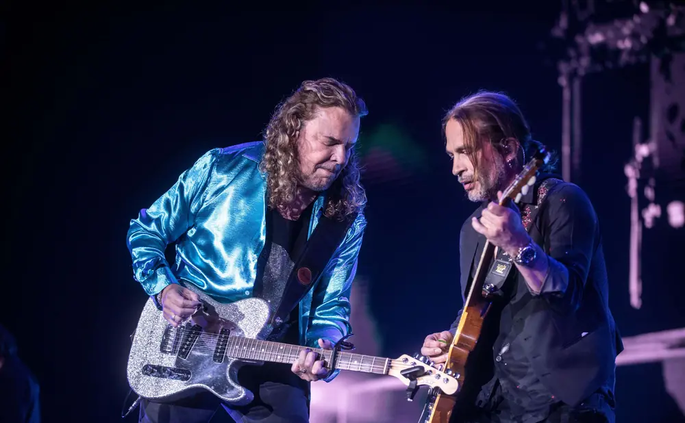 Two men playing guitars on stage. The man on the left wears a shiny blue jacket, and the man on the right wears a dark suit jacket.