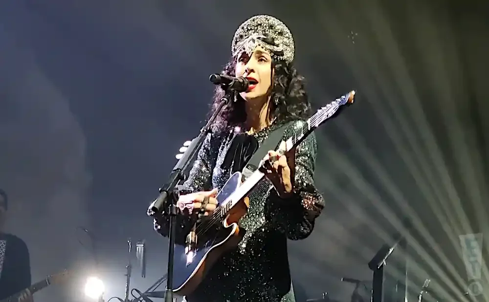 Singer with elaborate headdress and sparkly outfit plays guitar on stage.