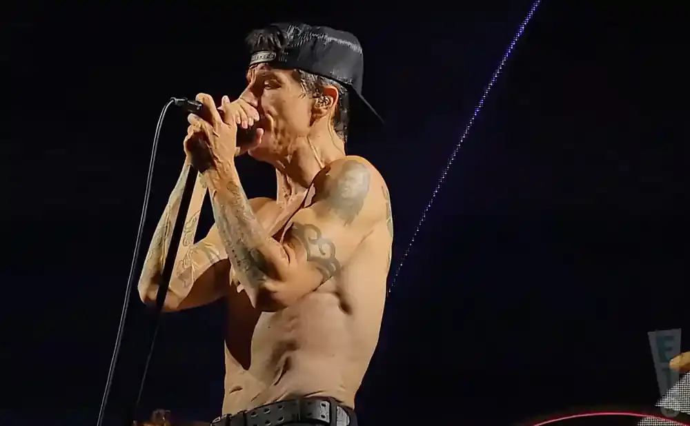 A shirtless man in a black baseball cap sings into a microphone on stage.